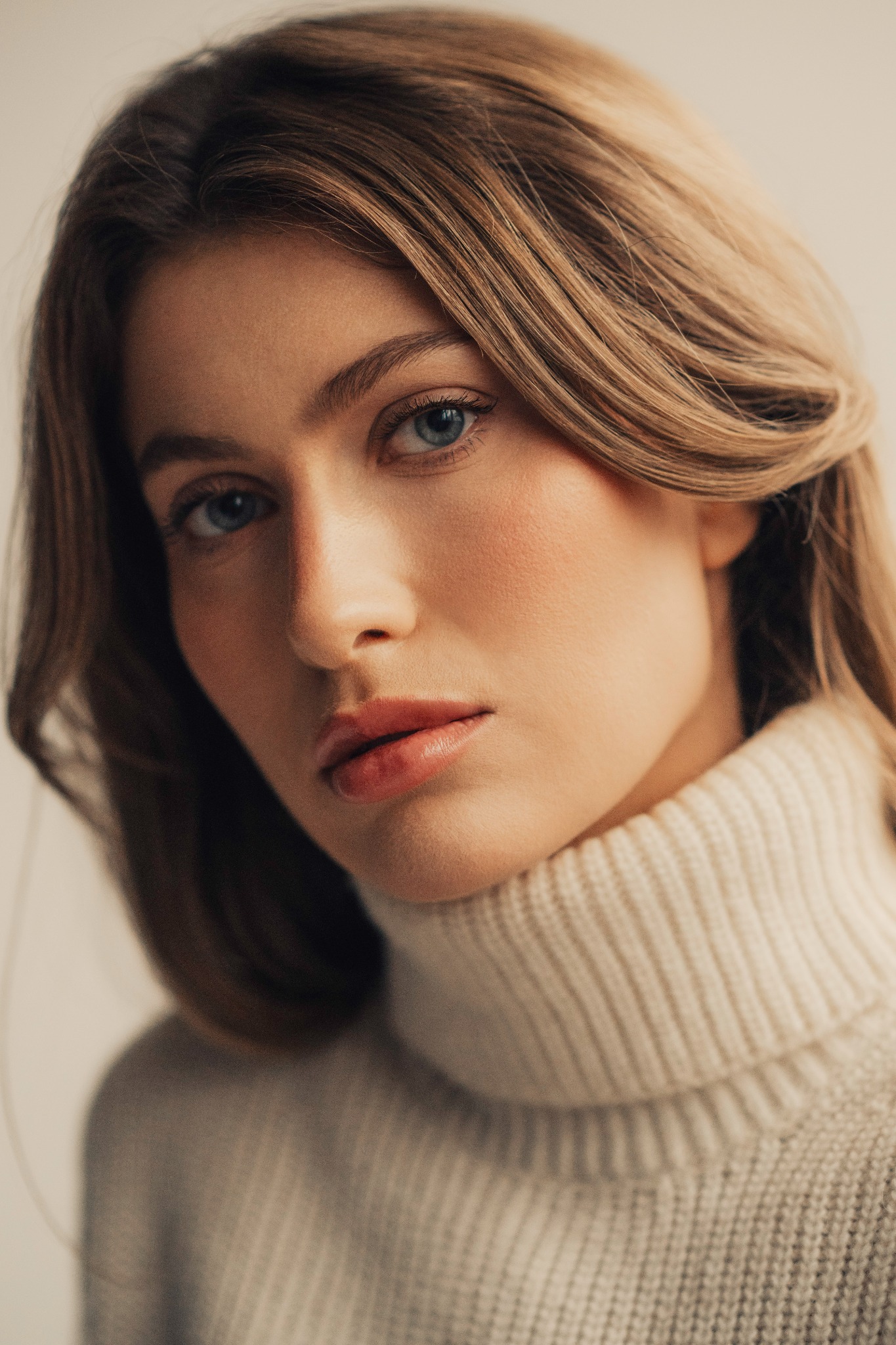 Portret młodej kobiety o jasnych włosach i niebieskich oczach, ubrana w kremowy golf, delikatny makijaż, naturalne światło.