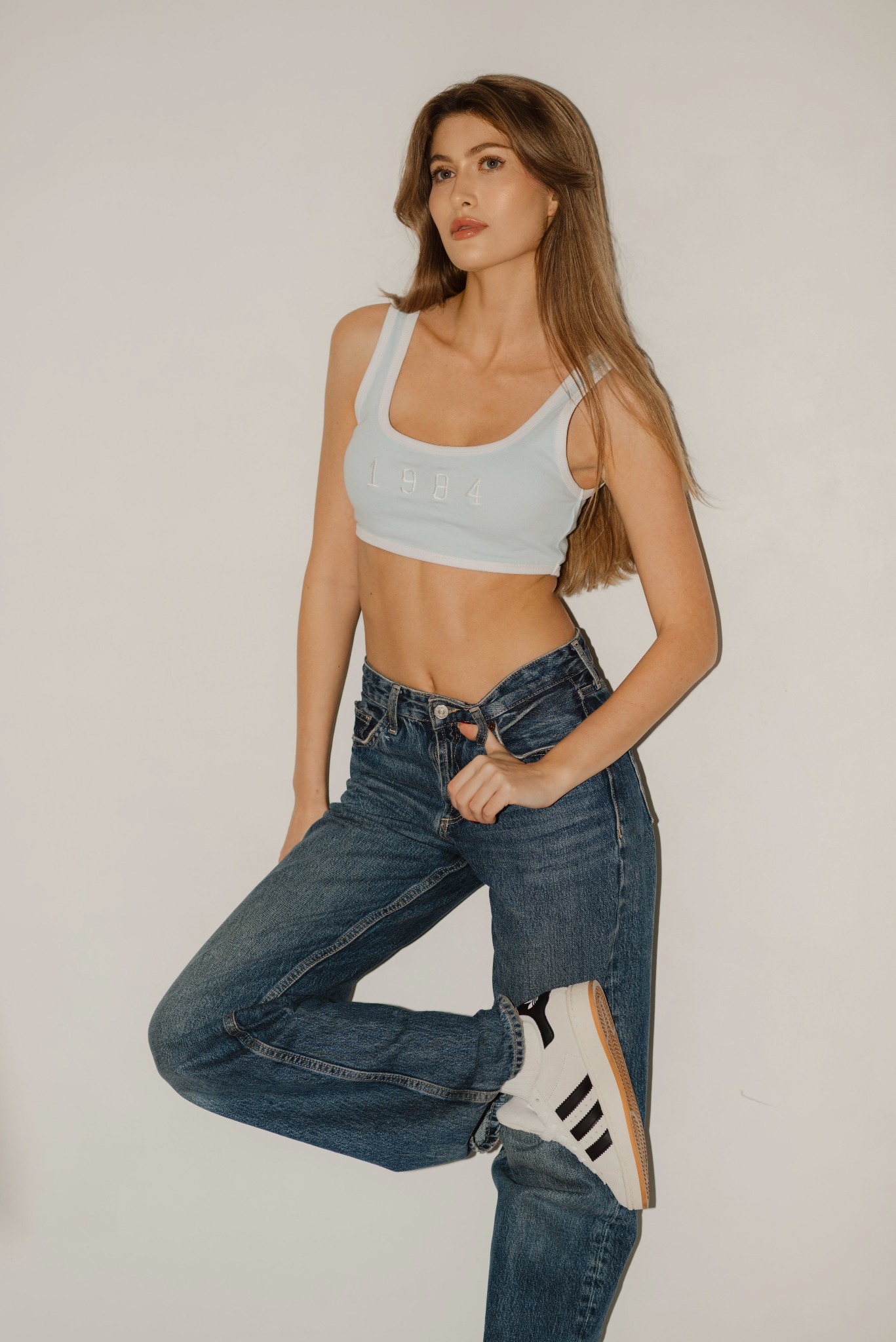 Portret kobiety w dżinsach i topie sportowym z napisem '1984', pozującej na białym tle z uniesioną nogą i butem sportowym z trzema paskami.