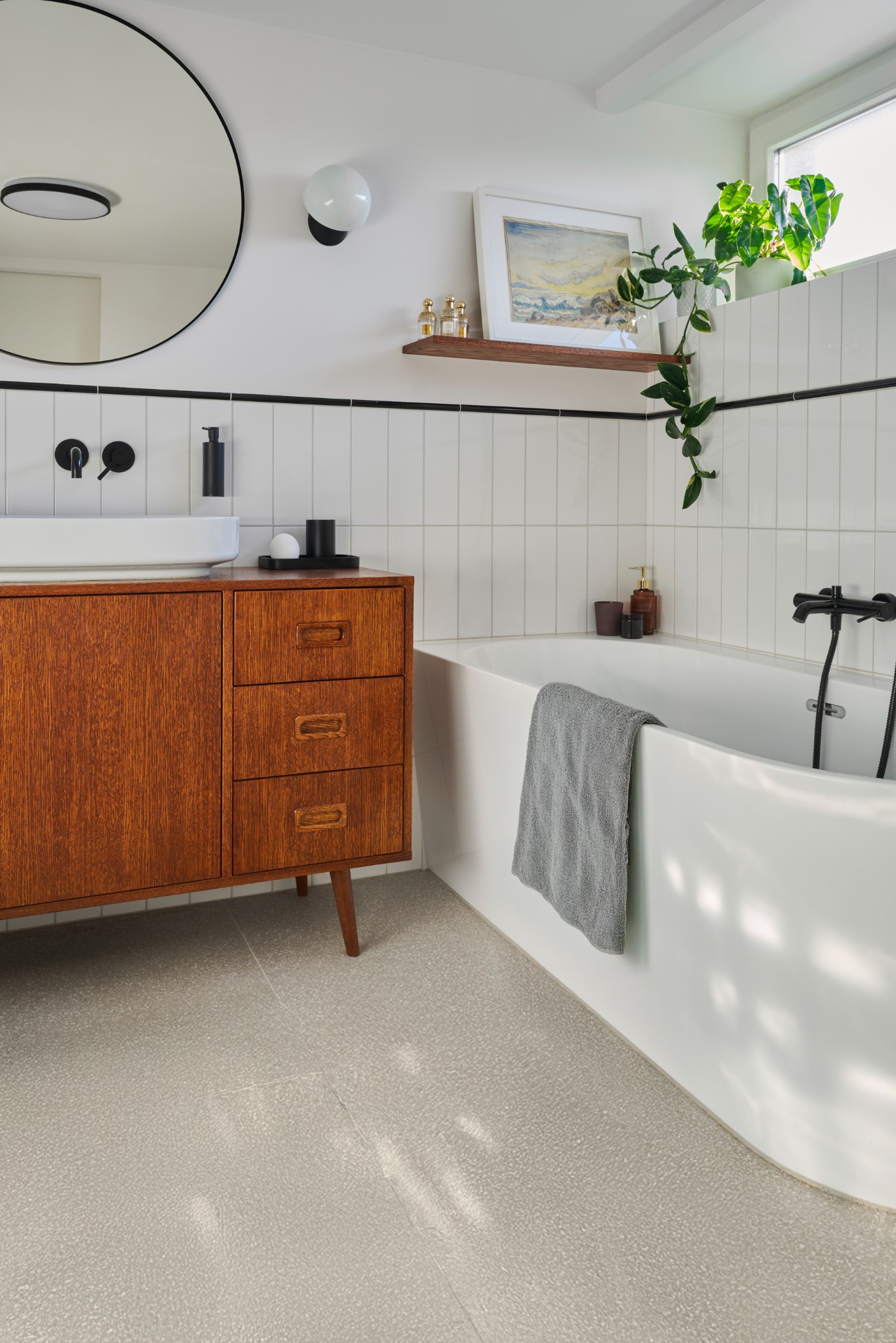 Nowoczesna łazienka z drewnianą szafką pod umywalką, białą wanną z czarną armaturą, jasnymi płytkami i okrągłym lustrem. Na półce nad wanną dekoracje i roślina.