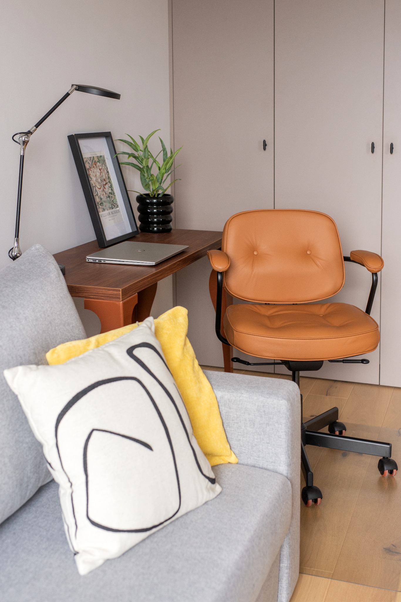 Nowoczesne biuro domowe z brązowym skórzanym krzesłem biurowym, drewnianym biurkiem z laptopem, lampą i rośliną w czarnej doniczce, obok szarej sofy z żółtą i białą poduszką z czarnym wzorem.