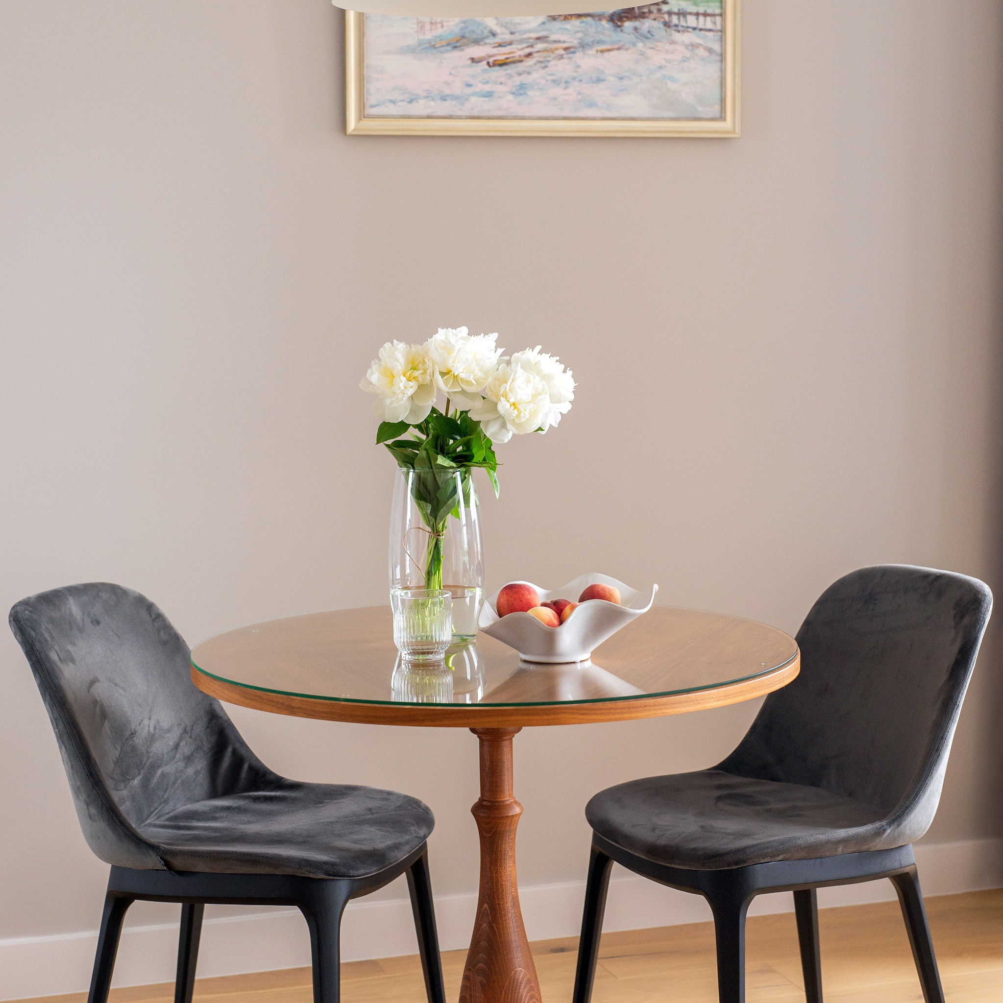Aranżacja wnętrza: okrągły stół z szklanym blatem, bukiet białych piwonii w wazonie, owoce w ceramicznej misie, dwie szare krzesła z welurowym obiciem, obraz na ścianie.