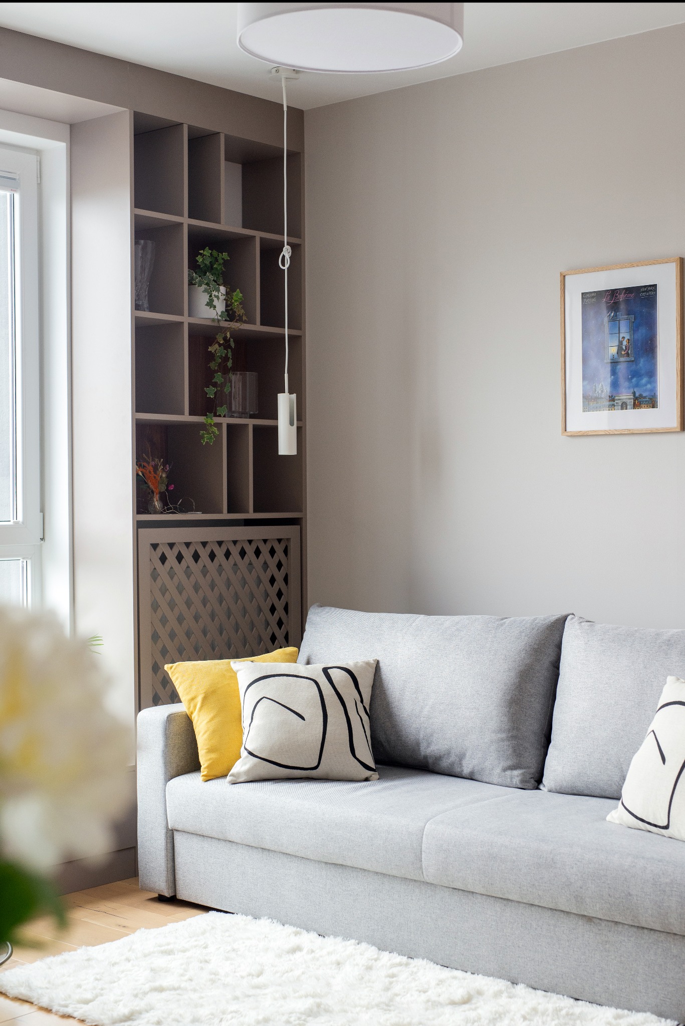 Jasnoszara sofa z żółtą i wzorzystymi poduszkami, drewniany regał z roślinami, obraz na ścianie i puszysty dywan w minimalistycznym salonie.