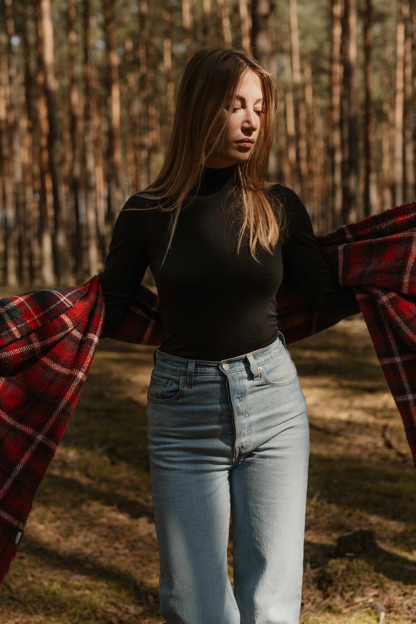 Portret kobiety w czarnym golfie i dżinsach, na tle leśnych drzew, z zarzuconym na ramiona czerwono-czarnym pledem w kratę, w naturalnym świetle.