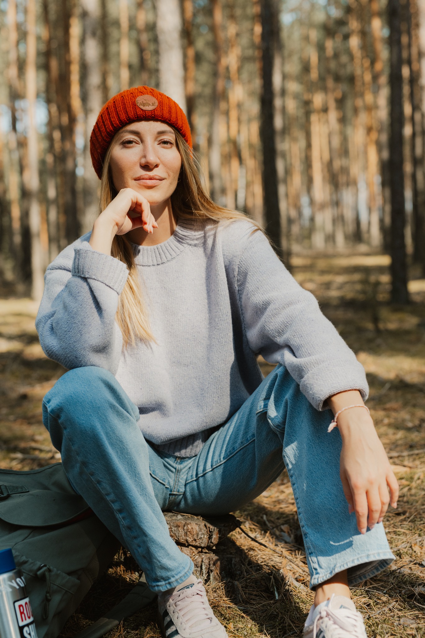 Portret kobiety w pomarańczowej czapce siedzącej na pniu drzewa w lesie, ubrana w jasny sweter i dżinsy, z plecakiem i butelką obok.