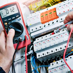 Elektryk Poznań - ELEKTRO-PROGRES - Instalacje Elektryczne - Elektryk w kasku, mierzący napięcie w skrzynce rozdzielczej za pomocą multimetru cyfrowego z czerwonymi przewodami, widoczne bezpieczniki i okablowanie.