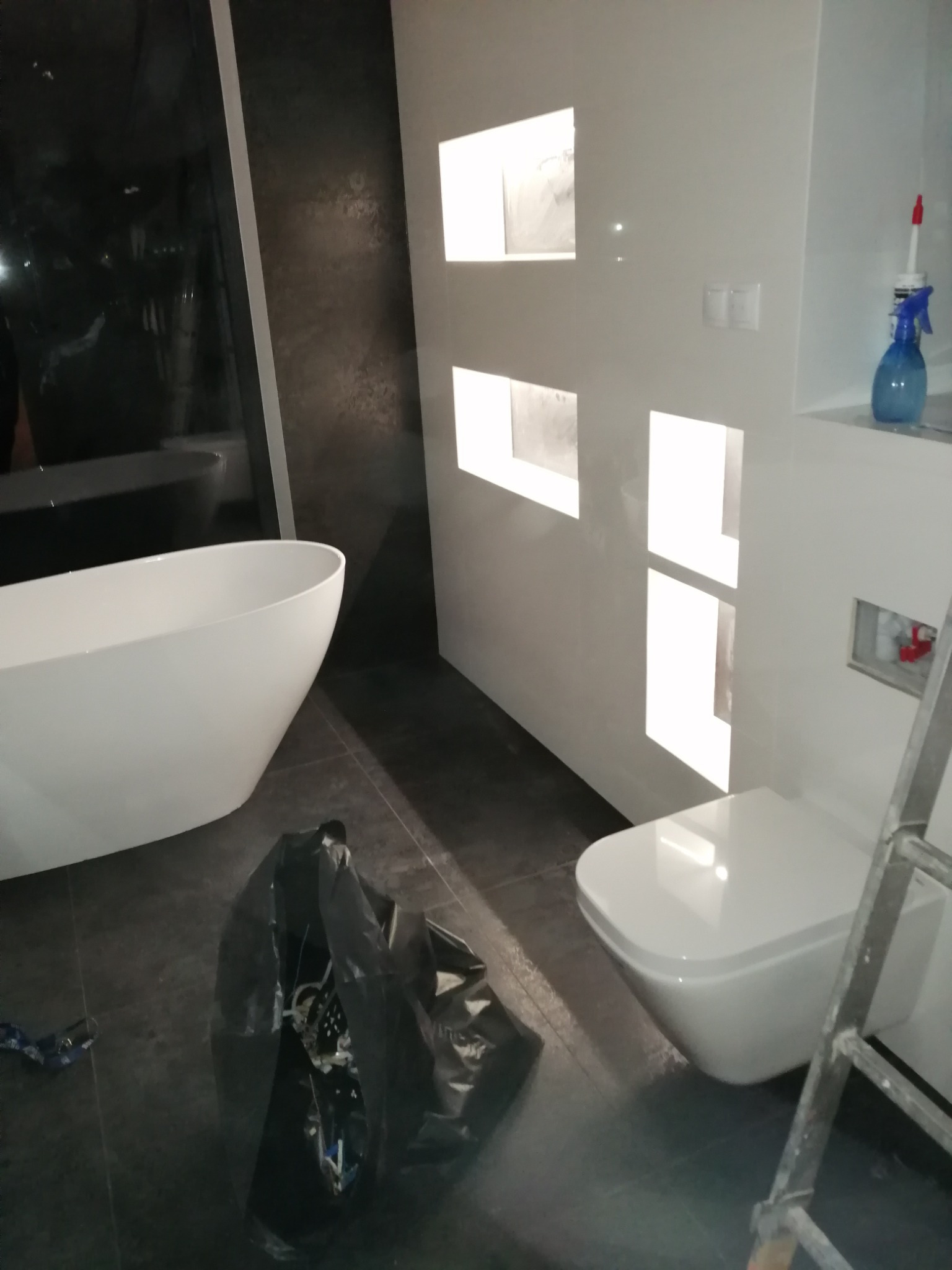 Nowoczesna łazienka w trakcie wykańczania: biała wolnostojąca wanna, podwieszana toaleta, białe ściany z dekoracyjnymi wnękami, ciemna podłoga, widoczna drabina malarska i worek z narzędziami.