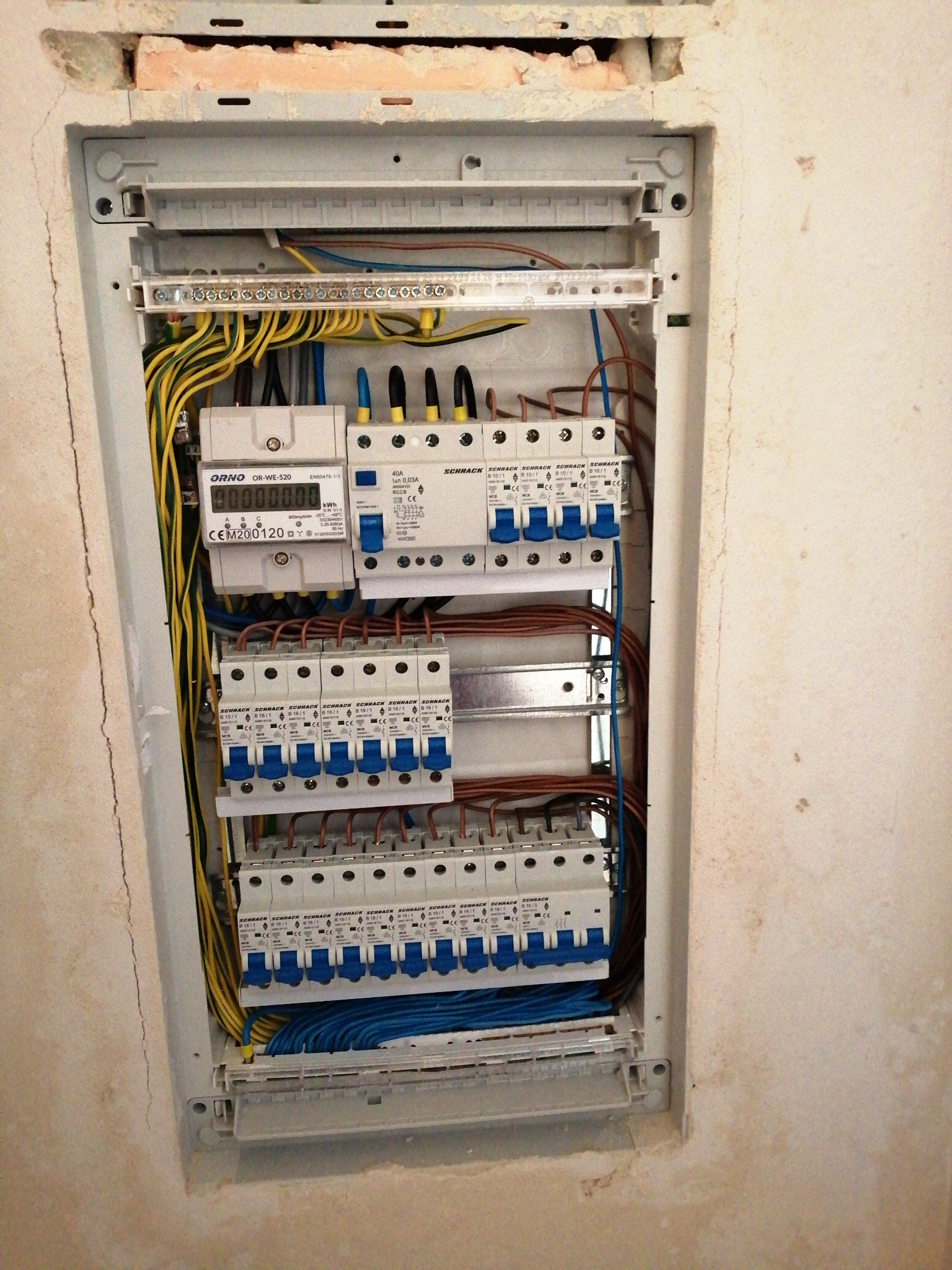 Rozdzielnica elektryczna wnękowa, z zamontowanymi bezpiecznikami, licznikiem ORNO i przewodami w kolorach żółtym, niebieskim i brązowym, widoczne niedoskonałości ściany wokół wnęki.