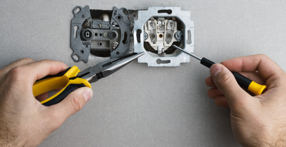 Elektryk przy pracy: demontaż gniazdka elektrycznego za pomocą kombinerek z żółtymi uchwytami i śrubokręta z czarno-żółtą rączką na tle szarej ściany.