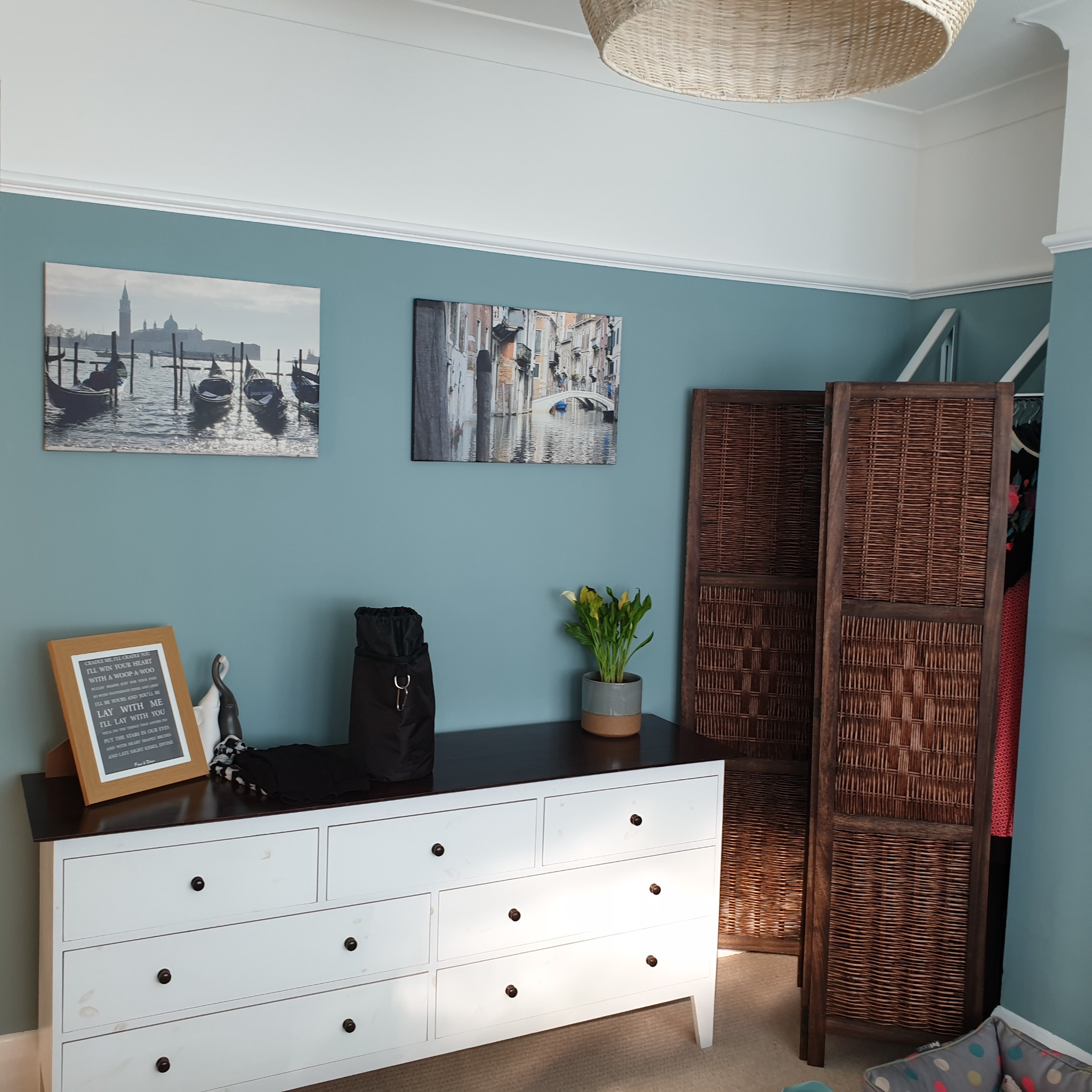 Pokój z turkusowymi ścianami, białymi listwami przypodłogowymi, komodą z ciemnym blatem, dekoracjami na ścianach i parawanem z wikliny.