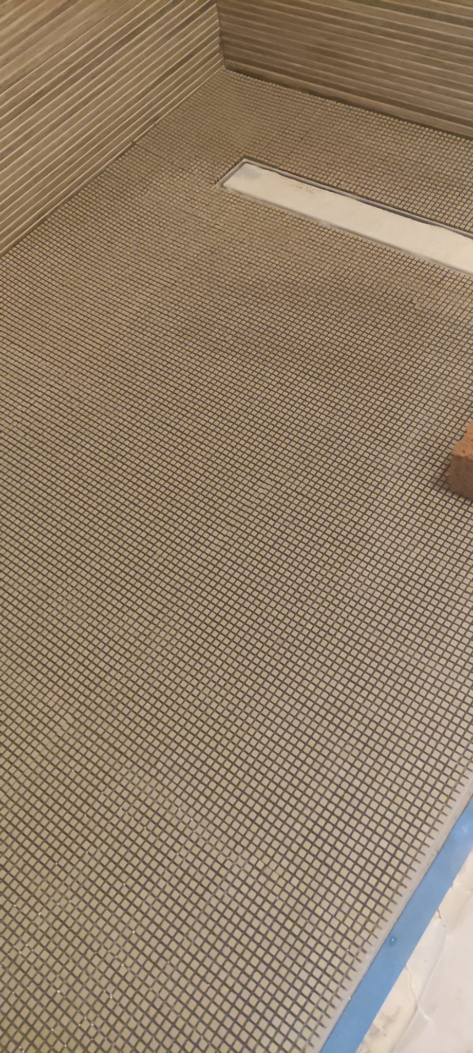 Przygotowana powierzchnia podłoża w łazience z odpływem liniowym, pokryta siatką wzmacniającą przed położeniem płytek, widoczny fragment cegły.