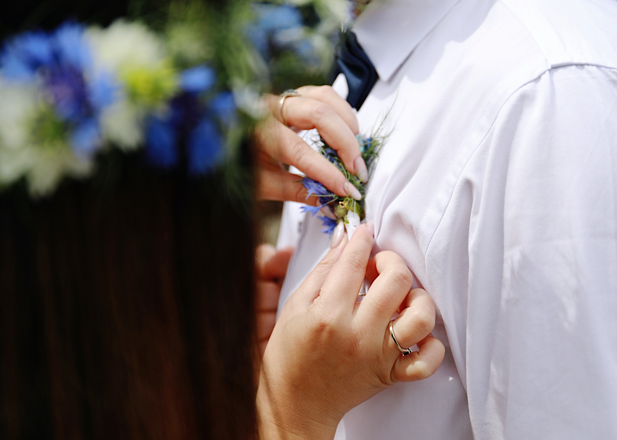 Przypinanie butonierki z chabrami do białej koszuli pana młodego, widoczne dłonie z pierścionkami i fragment wianka z błękitnych kwiatów.
