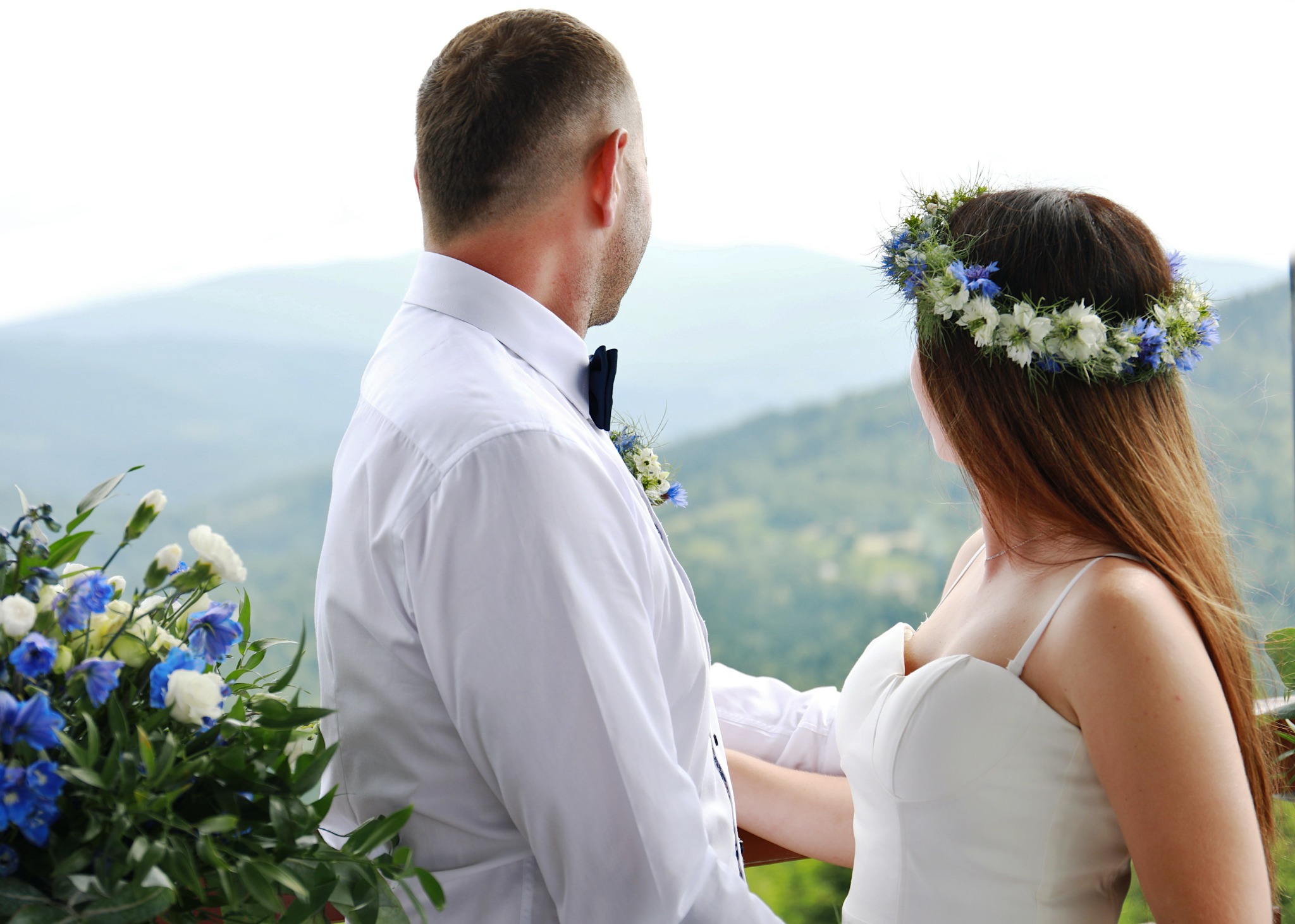 Para młoda, on w białej koszuli i muszce, ona w białej sukni i wianku z białych i niebieskich kwiatów, stoi na tle górskiego krajobrazu.