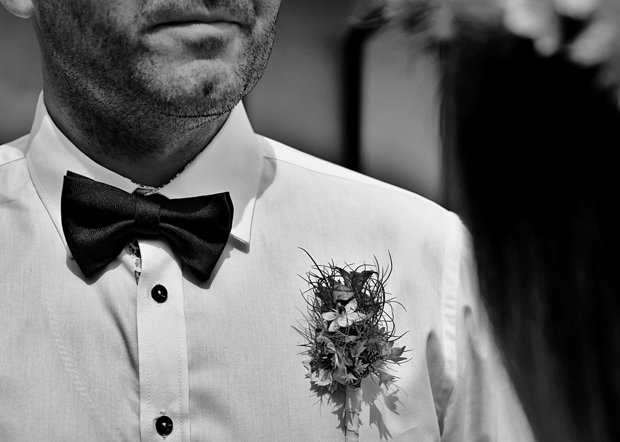 Czarno-białe zbliżenie mężczyzny w białej koszuli z czarną muchą i oryginalną butonierką z drobnych kwiatów i gałązek.
