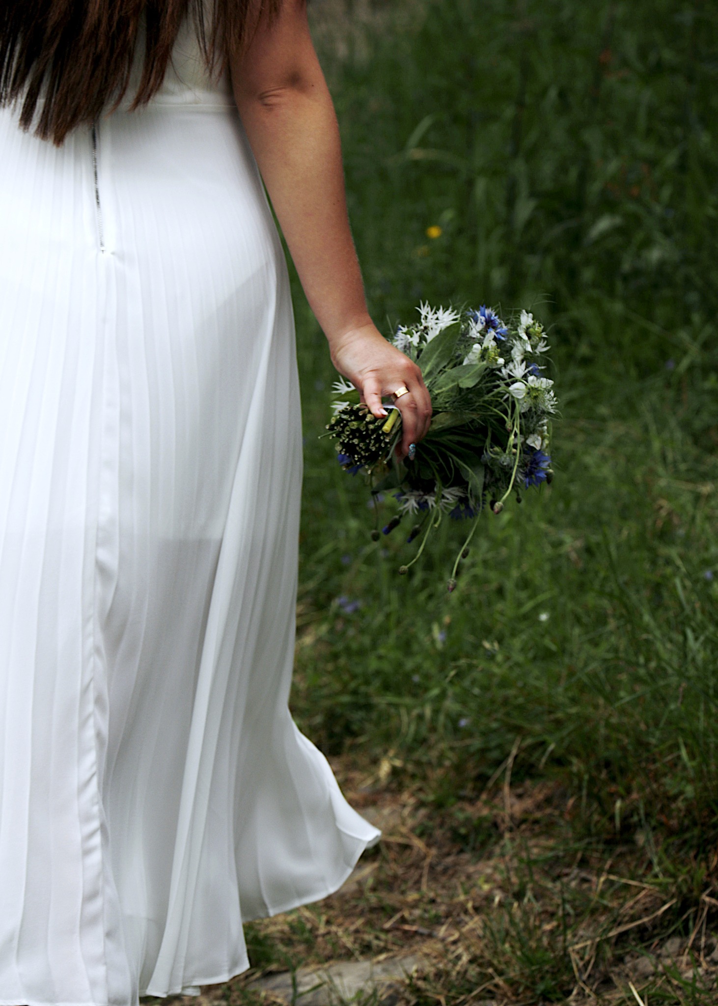 Panna młoda w białej, plisowanej sukni trzyma bukiet polnych kwiatów z niebieskimi chabrami i białymi margaretkami, widoczny złoty pierścionek na palcu.