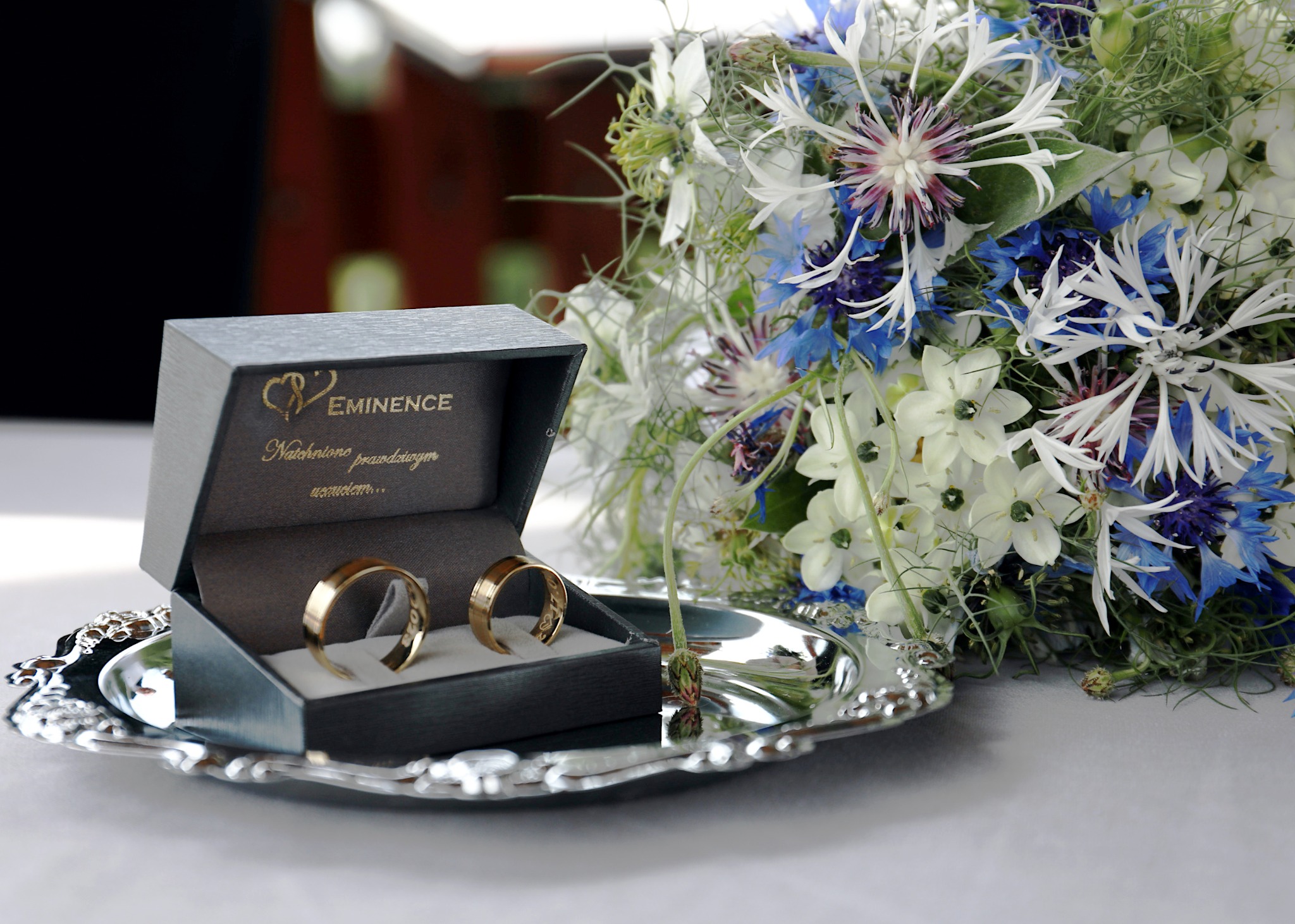 Dwa złote obrączki w szarym pudełku marki Eminence, ustawione na srebrnej tacy obok bukietu z białych i niebieskich kwiatów.