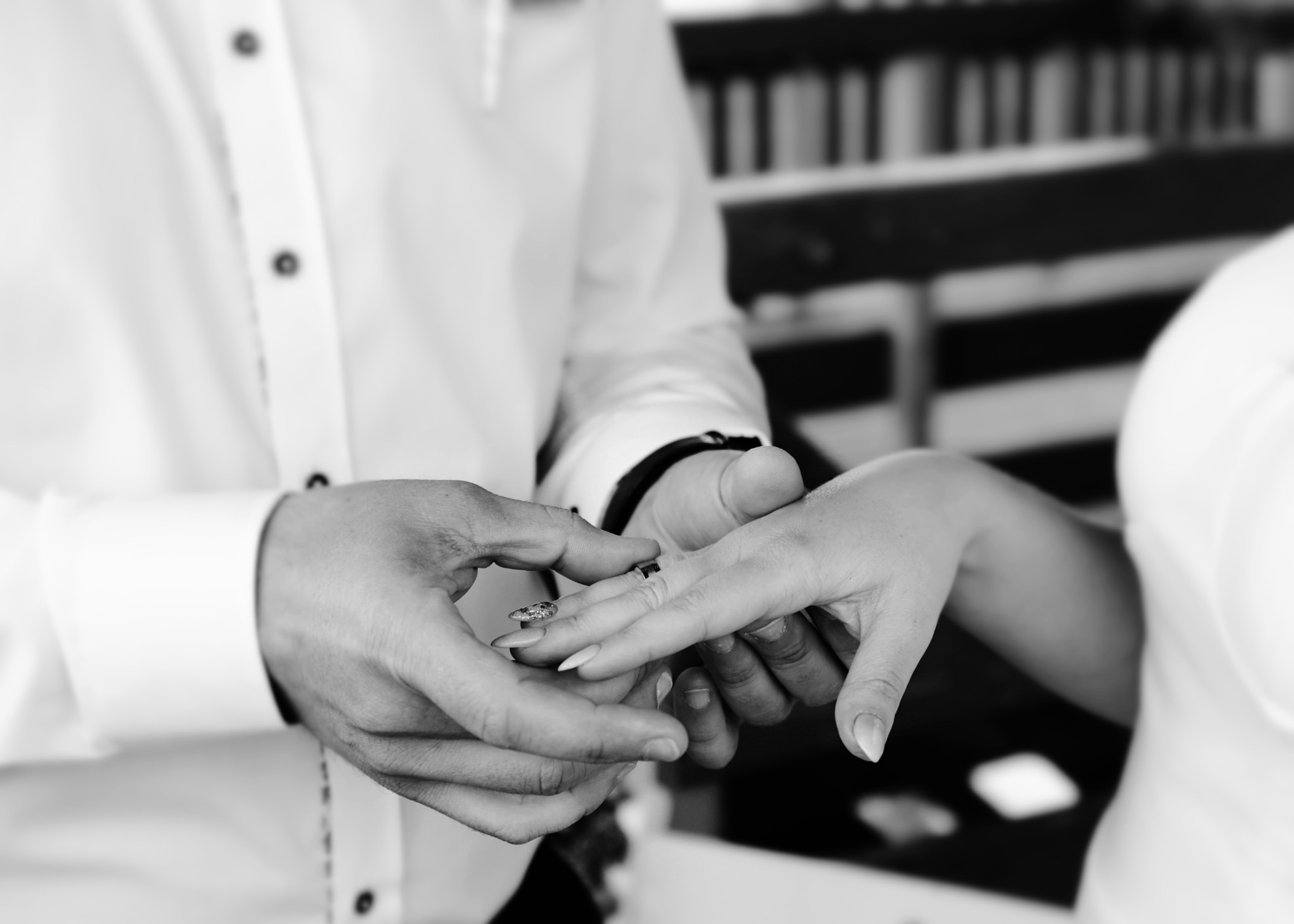 Czarno-białe zbliżenie dłoni zakładających obrączkę ślubną na palec panny młodej, widoczna mankiet koszuli pana młodego i delikatne zdobienie paznokci panny młodej.