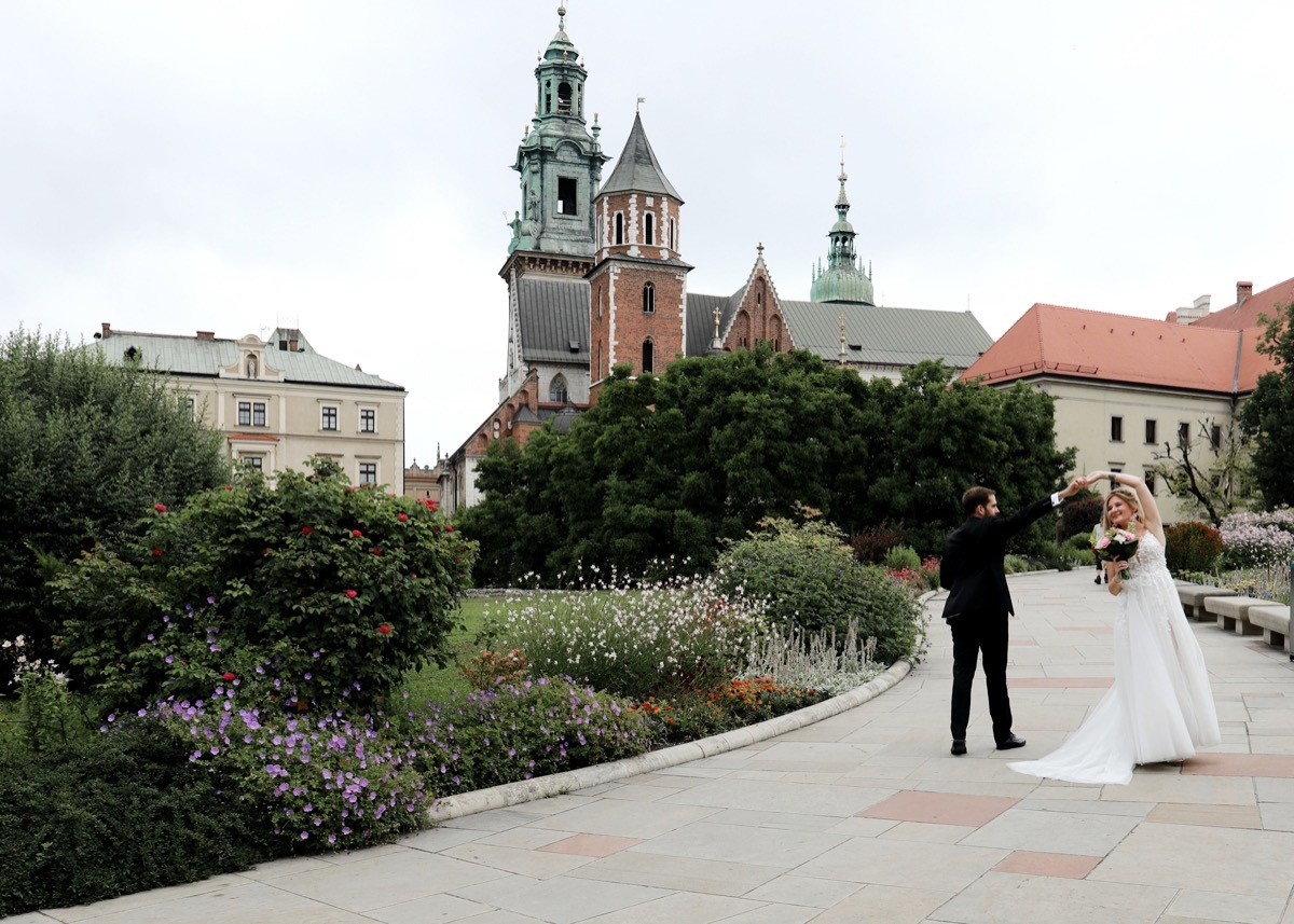 Para młoda w tańcu na tle Wawelu w Krakowie, panna młoda w białej sukni z bukietem, pan młody w czarnym garniturze, ujęcie z dołu, kwiaty na pierwszym planie.