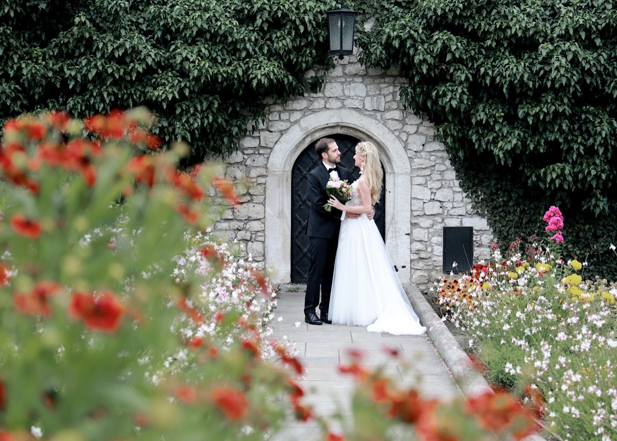 Para młoda w objęciach stoi przed kamiennym wejściem porośniętym bluszczem; w tle kwiaty i latarnia.