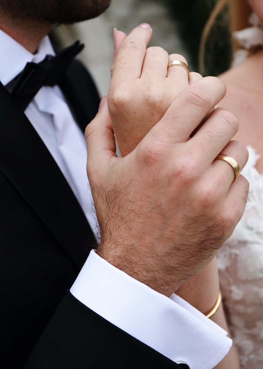 Dłonie nowożeńców splecione razem, widoczne złote obrączki na palcach, elegancki czarny smoking z białą koszulą i mankietami oraz fragment koronkowej sukni ślubnej.