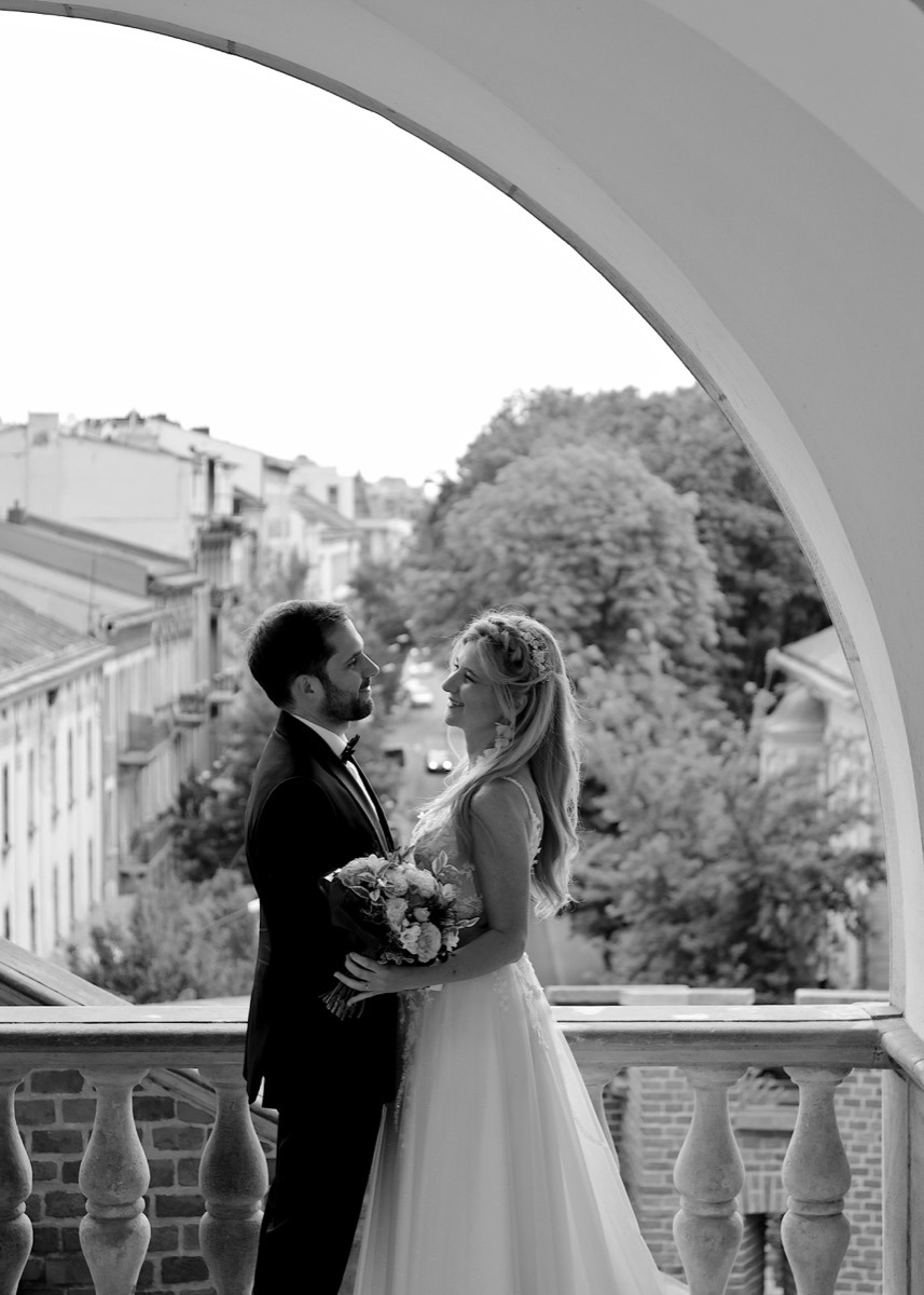 Czarno-białe zdjęcie pary młodej na balkonie z widokiem na miasto. Panna młoda trzyma bukiet kwiatów i patrzy na pana młodego.