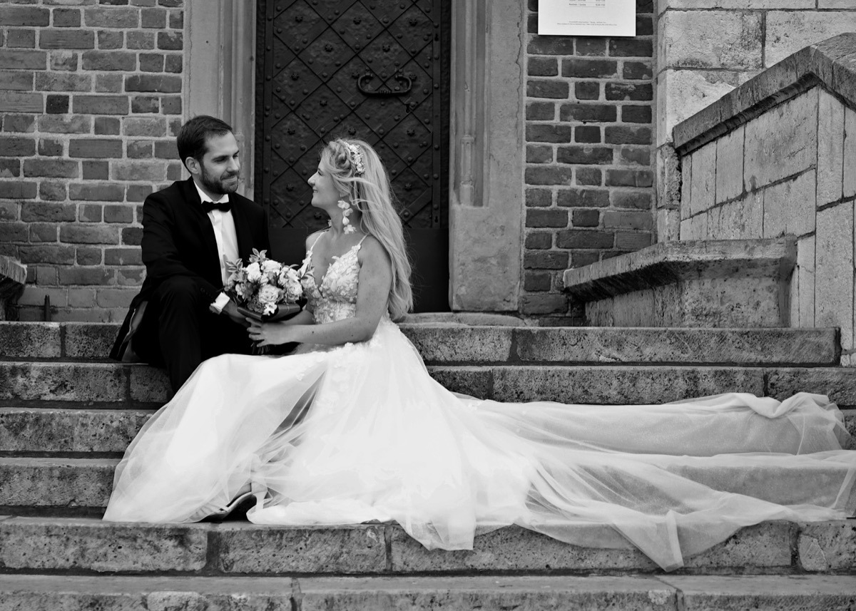 Czarno-białe zdjęcie pary młodej siedzącej na kamiennych schodach na tle ceglanej ściany i ciemnych drzwi. Panna młoda w sukni z rozcięciem trzyma bukiet, a pan młody w smokingu patrzy na nią...