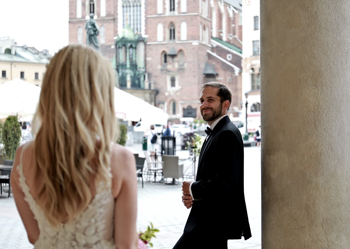 Pan młody w czarnym smokingu spogląda z uśmiechem na pannę młodą w koronkowej sukni ślubnej, w tle Rynek Główny w Krakowie.