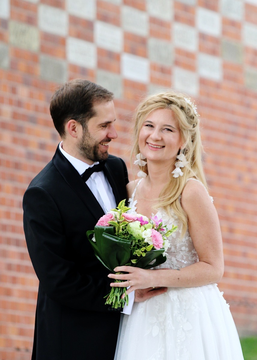 Uśmiechnięta para młoda w eleganckich strojach pozuje z bukietem na tle ceglanej ściany z szachownicą.