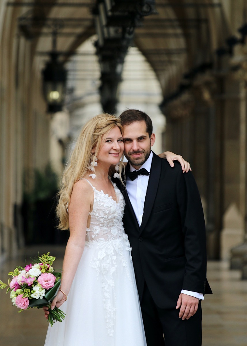 Uśmiechnięta para młoda w eleganckich strojach, ona w koronkowej sukni z bukietem, on w smokingu z muszką, pozuje na tle kolumnady z łukami i latarniami.
