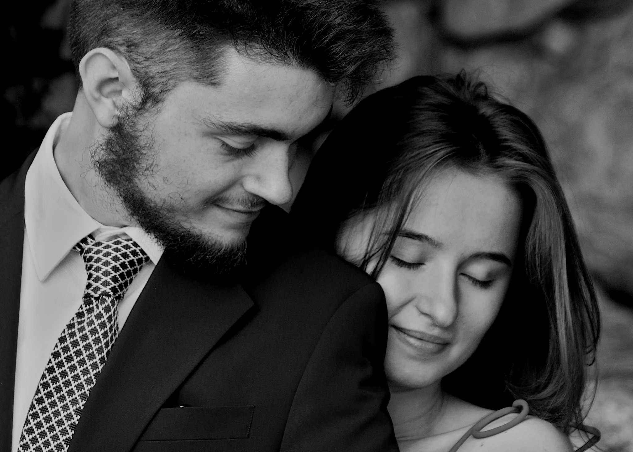 Czarno-białe zbliżenie pary: mężczyzna w garniturze i krawacie opiera głowę o głowę uśmiechniętej kobiety z zamkniętymi oczami.