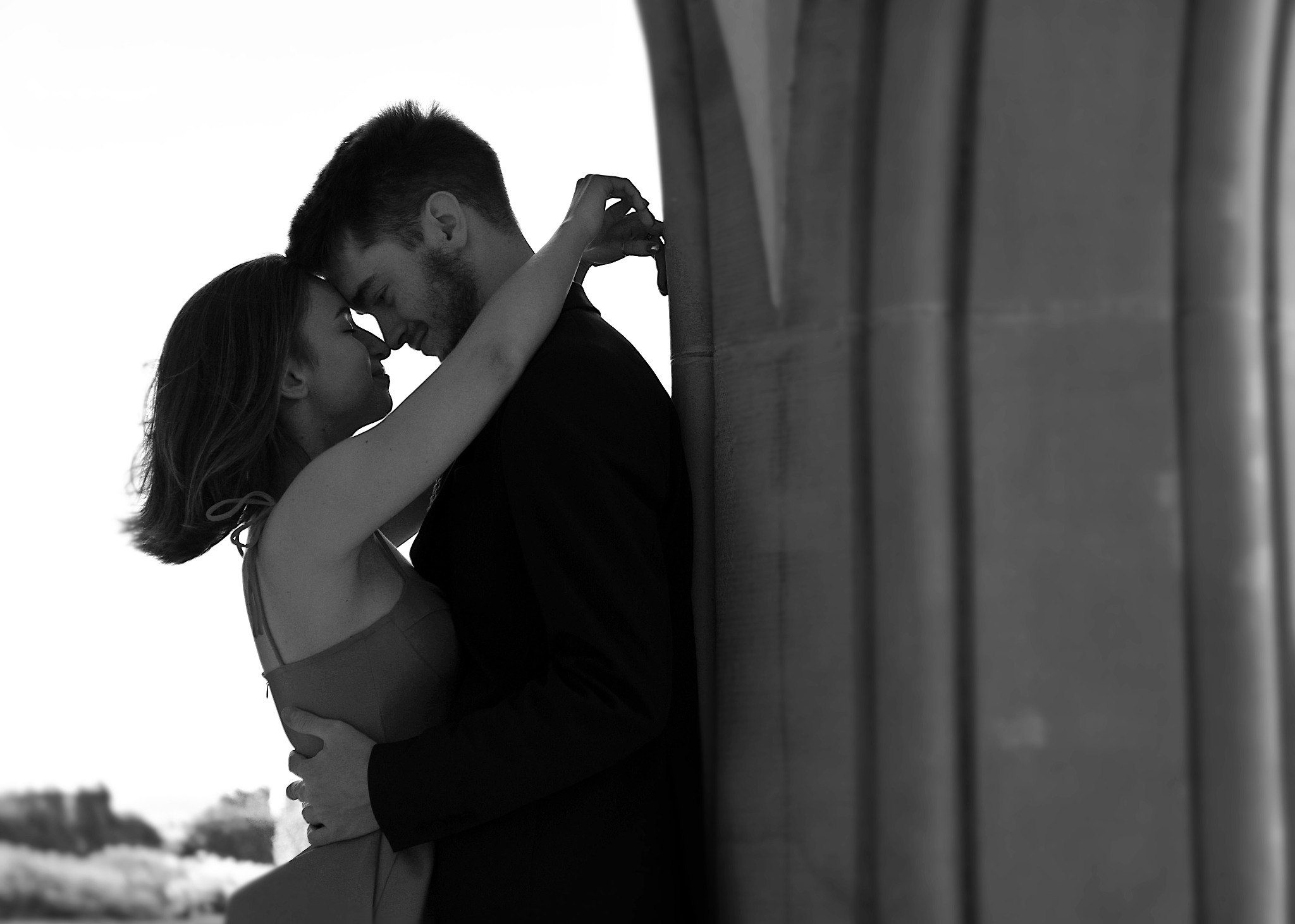 Czarno-białe zdjęcie zakochanej pary przytulającej się pod kamiennym łukiem, z widocznymi elementami architektury w tle.