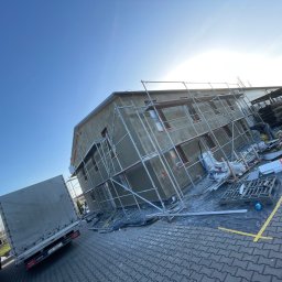 ComplexTerm - Budynek w trakcie tynkowania elewacji, widoczne rusztowania, materiały budowlane i pracownik w oddali, na pierwszym planie zaparkowane samochody dostawcze.