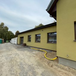 ComplexTerm - Budynek o żółtej elewacji z widocznym przyłączem wody i żółtym wężem na żwirowym podłożu, w tle zielony garaż blaszany i drzewa.