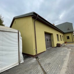 ComplexTerm - Żółty budynek z brązowymi elementami wykończeniowymi, widoczny z boku, z fragmentem białego namiotu po lewej stronie i brukowanym podjazdem, pochmurne niebo w tle.