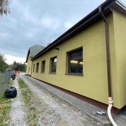 ComplexTerm - Żółta elewacja budynku z brązowymi oknami i rynnami, widok z boku, z workami na śmieci i wężem na żwirowej ścieżce obok.