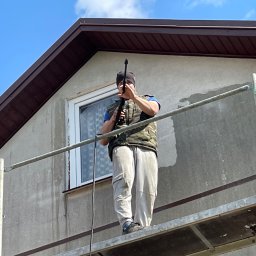 ComplexTerm - Pracownik na rusztowaniu czyści elewację domu za pomocą myjki ciśnieniowej, przygotowując ją do dalszych prac wykończeniowych.