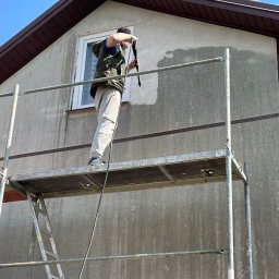 ComplexTerm - Pracownik na rusztowaniu czyści elewację domu myjką ciśnieniową, widoczny fragment wyczyszczonej ściany, pozostała część jest brudna.