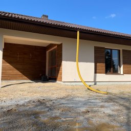 ComplexTerm - Dom jednorodzinny w trakcie prac elewacyjnych, widoczna żółta rura wentylacyjna, świeżo otynkowane ściany w jasnym odcieniu, brązowe okiennice i fragment drewnianej elewacji garażu, teren wokół...