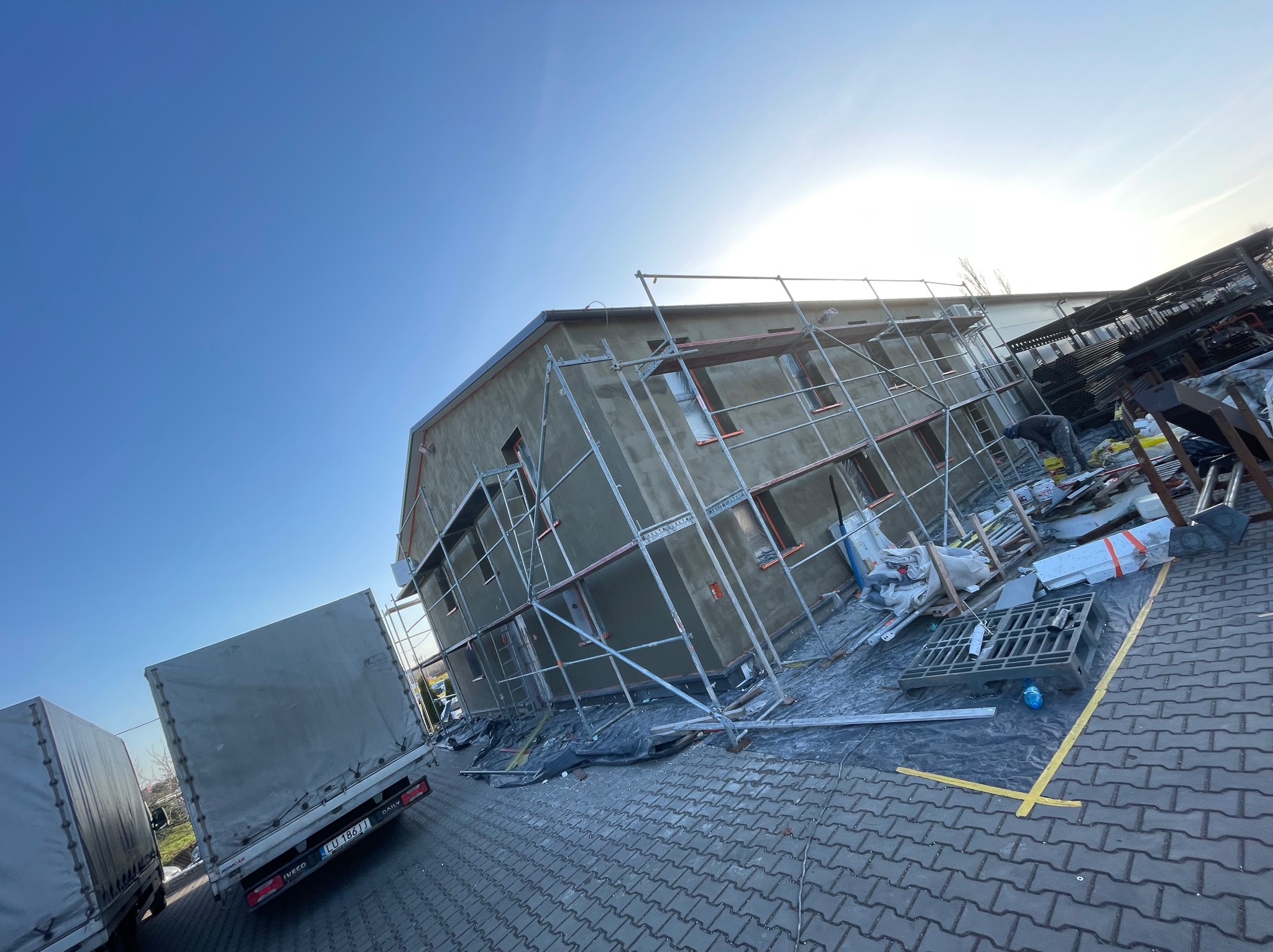 Budynek w trakcie tynkowania elewacji, widoczne rusztowania, materiały budowlane i pracownik w oddali, na pierwszym planie zaparkowane samochody dostawcze.