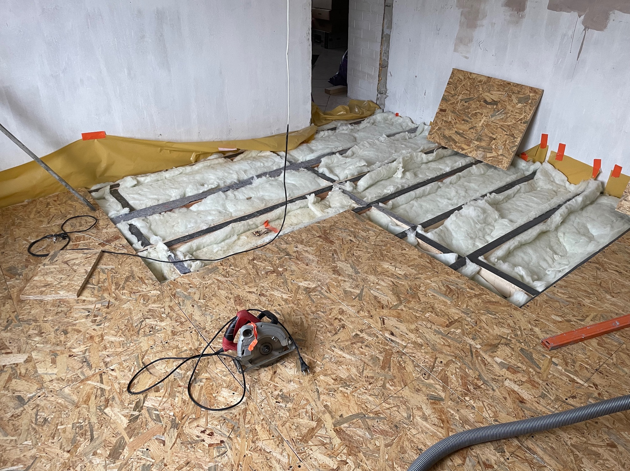 Wnętrze w trakcie prac: podłoga z płyt OSB z wyciętymi otworami, w których widać białą wełnę mineralną izolacyjną, żółta folia zabezpieczająca przy ścianie, piła elektryczna leżąca na podłodze.