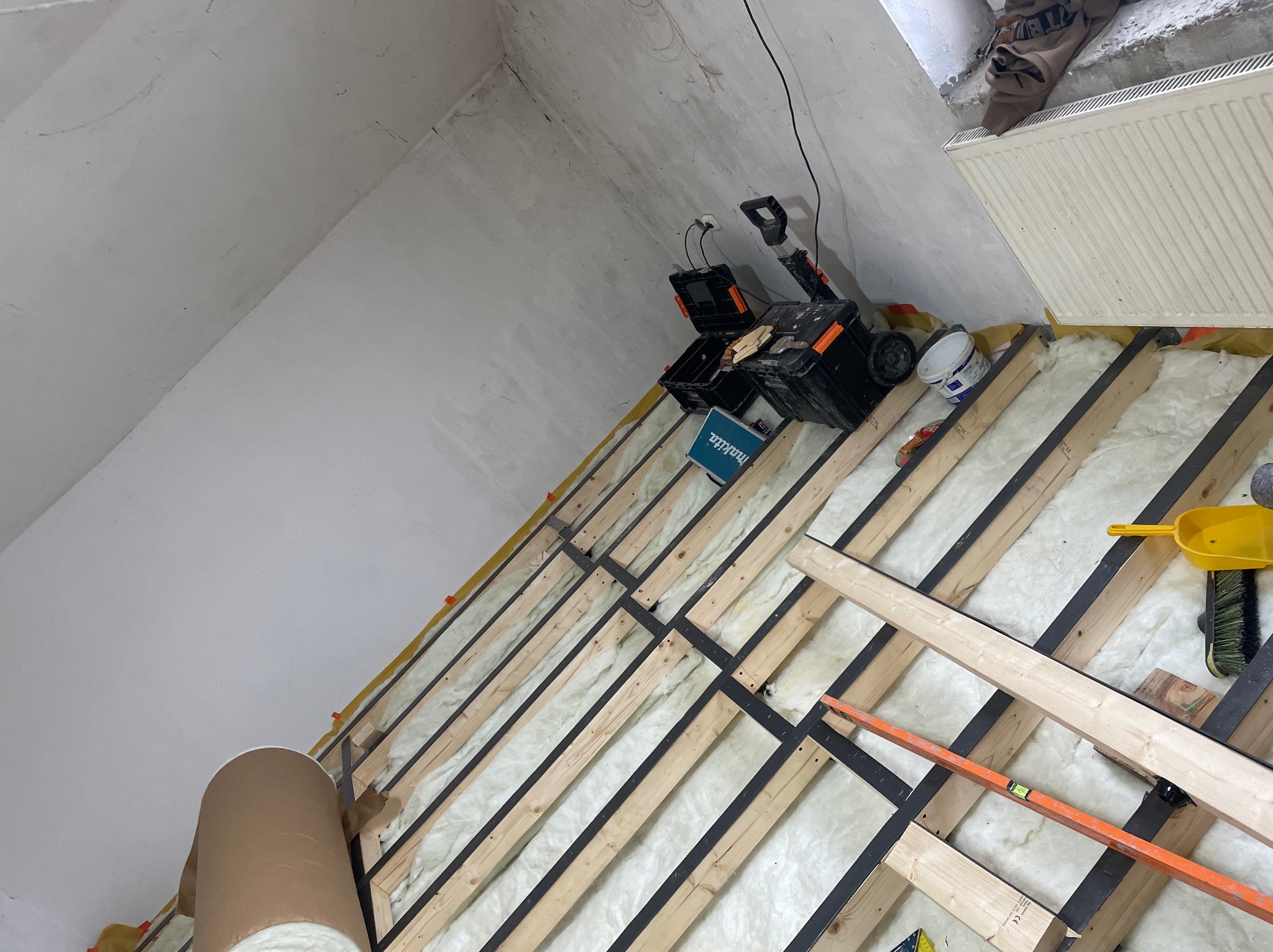 Skośny strych w trakcie izolacji wełną mineralną pomiędzy drewnianymi belkami, widoczne narzędzia, poziomica, wiadro z farbą i skrzynki narzędziowe.