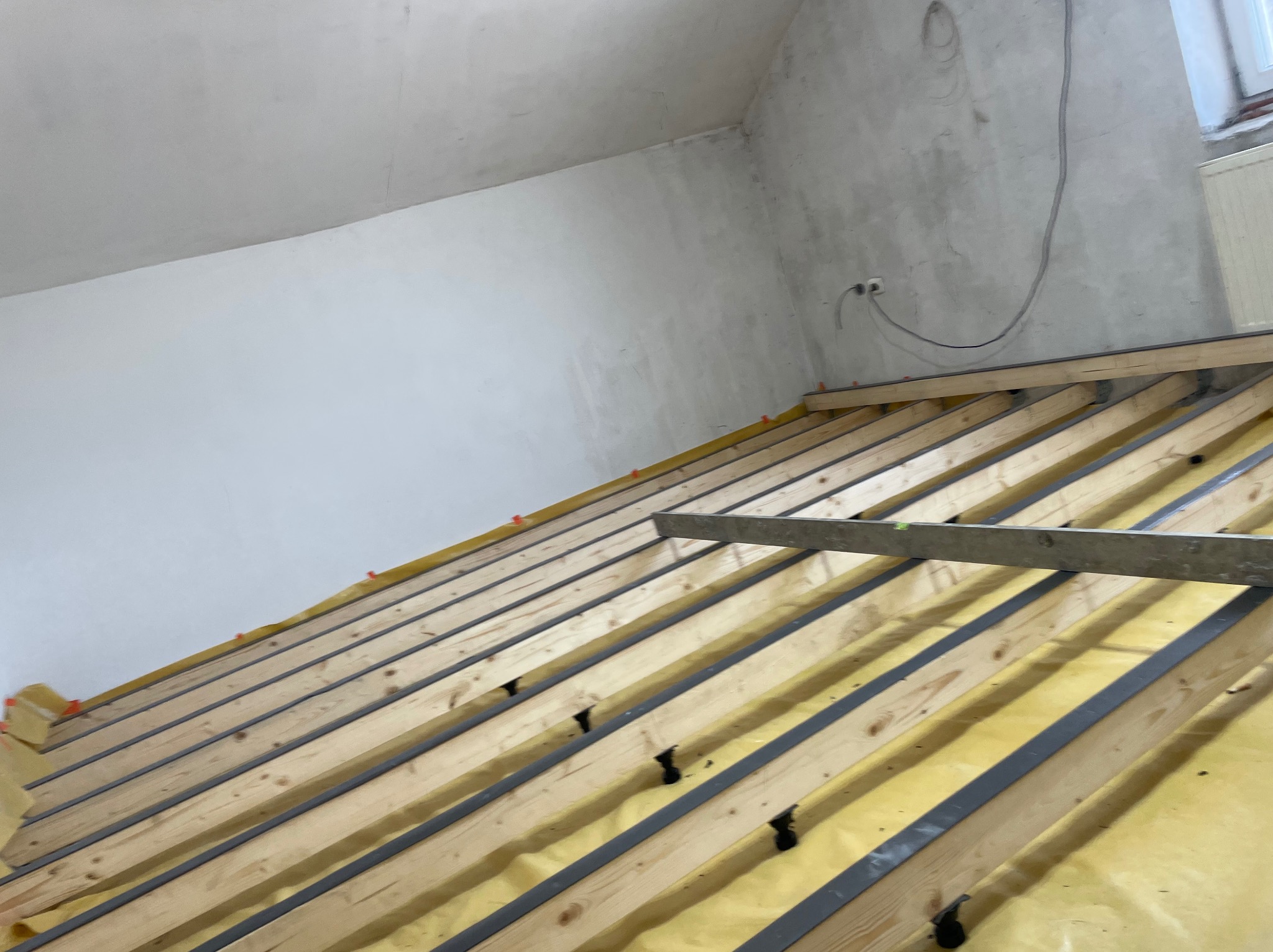 Wnętrze poddasza w trakcie prac: drewniana konstrukcja dachu z izolacją z folii w kolorze żółtym, widoczne listwy montażowe i poziomica, surowy stan ścian.