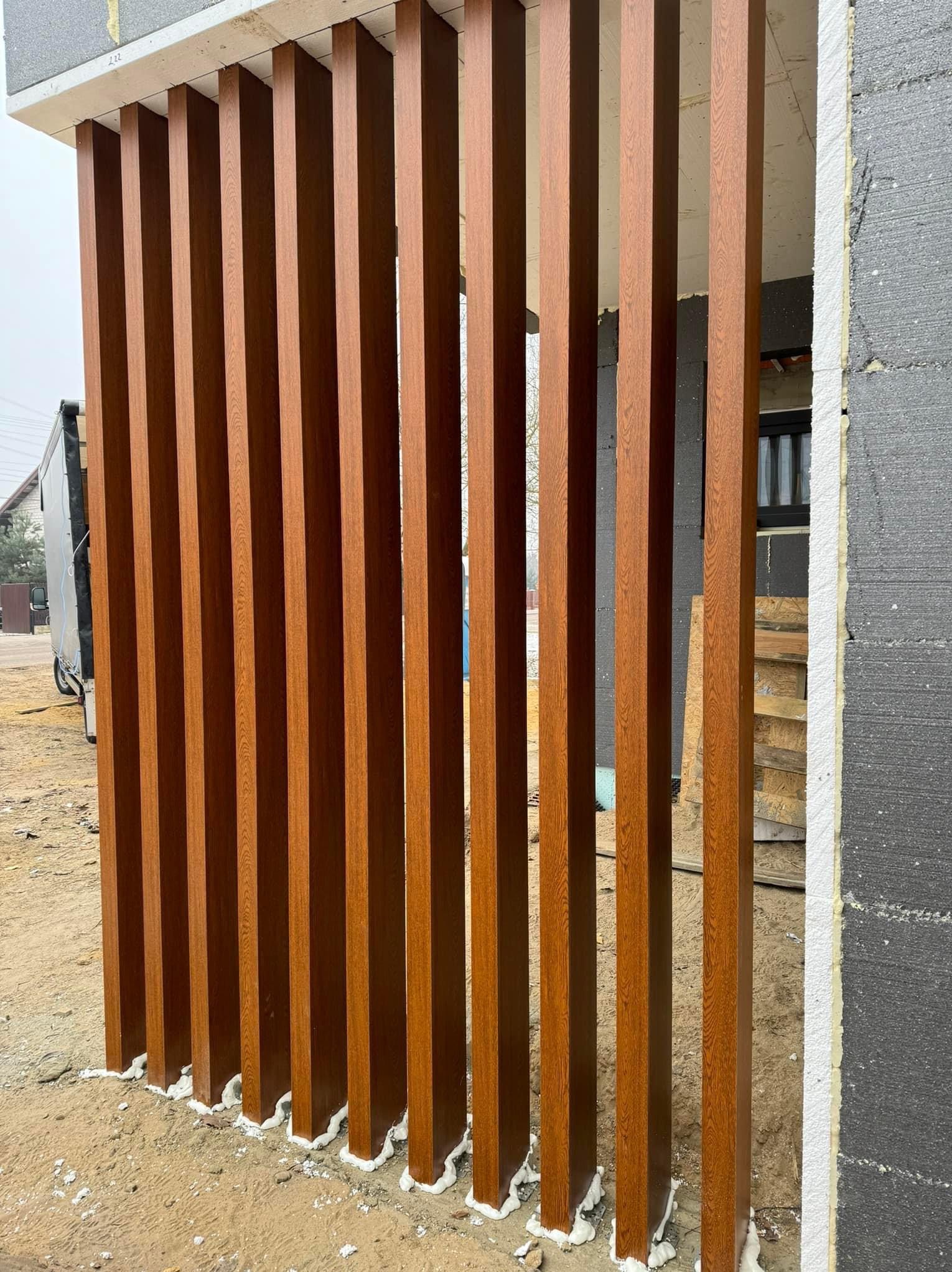 Drewnopodobne, pionowe panele elewacyjne zamontowane na budynku z ociepleniem, widoczne szpary między panelami i pianka montażowa u podstawy.