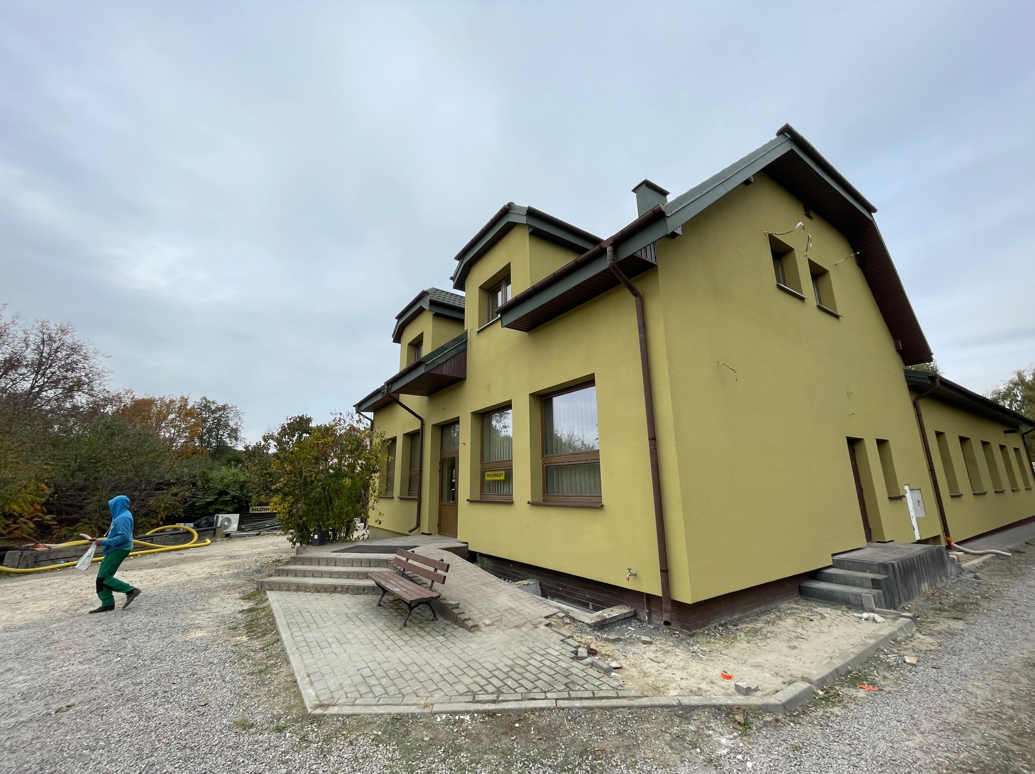 Budynek po tynkowaniu elewacji na kolor oliwkowy, widoczny fragment chodnika z kostki brukowej i osoba w ubraniu roboczym.