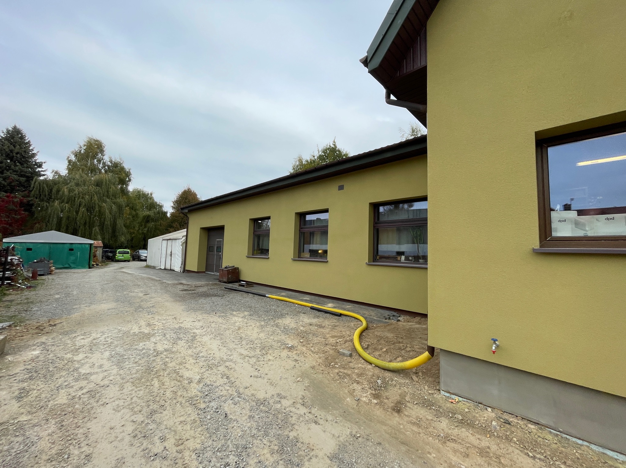 Budynek o żółtej elewacji z widocznym przyłączem wody i żółtym wężem na żwirowym podłożu, w tle zielony garaż blaszany i drzewa.
