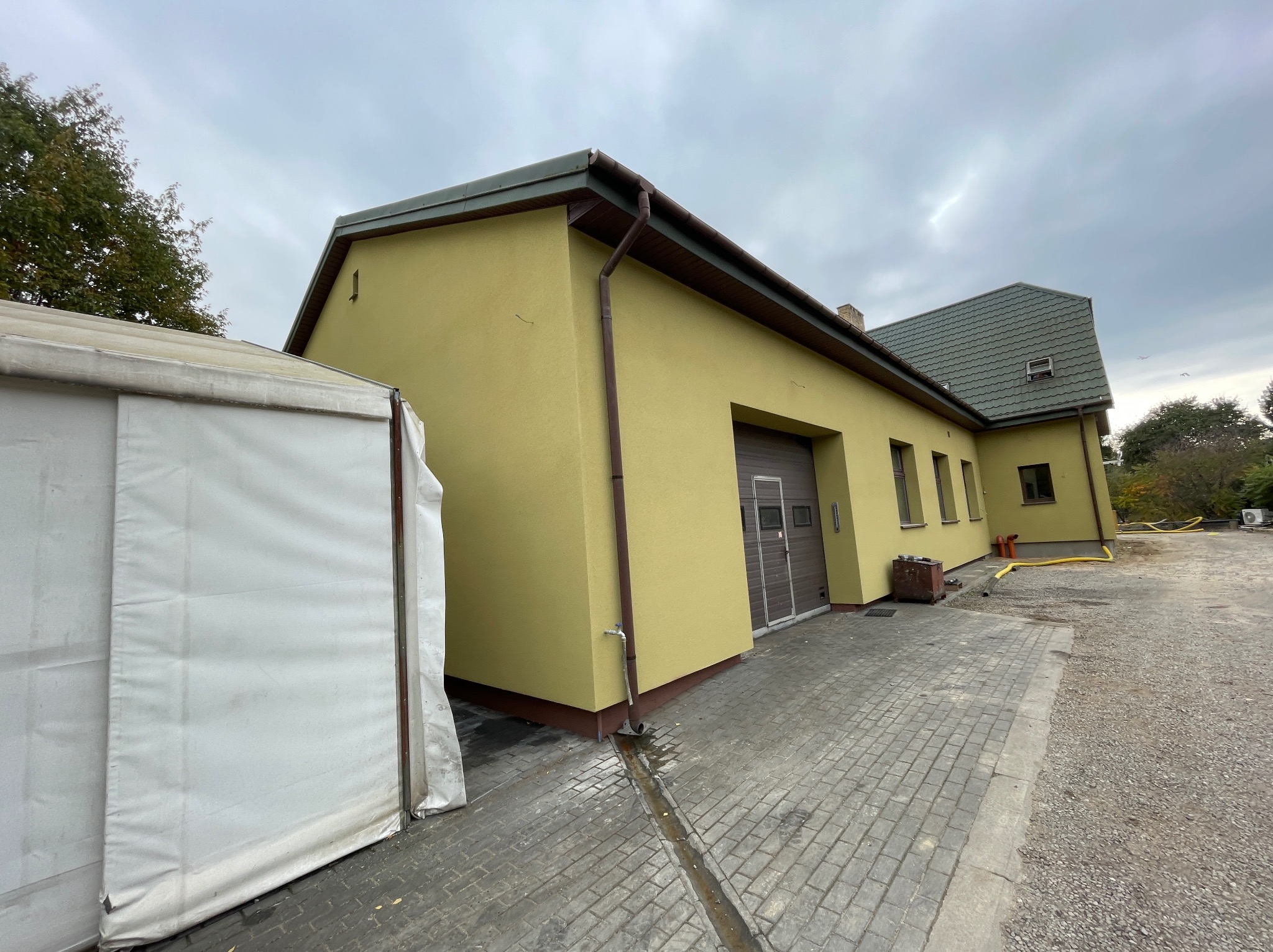Żółty budynek z brązowymi elementami wykończeniowymi, widoczny z boku, z fragmentem białego namiotu po lewej stronie i brukowanym podjazdem, pochmurne niebo w tle.