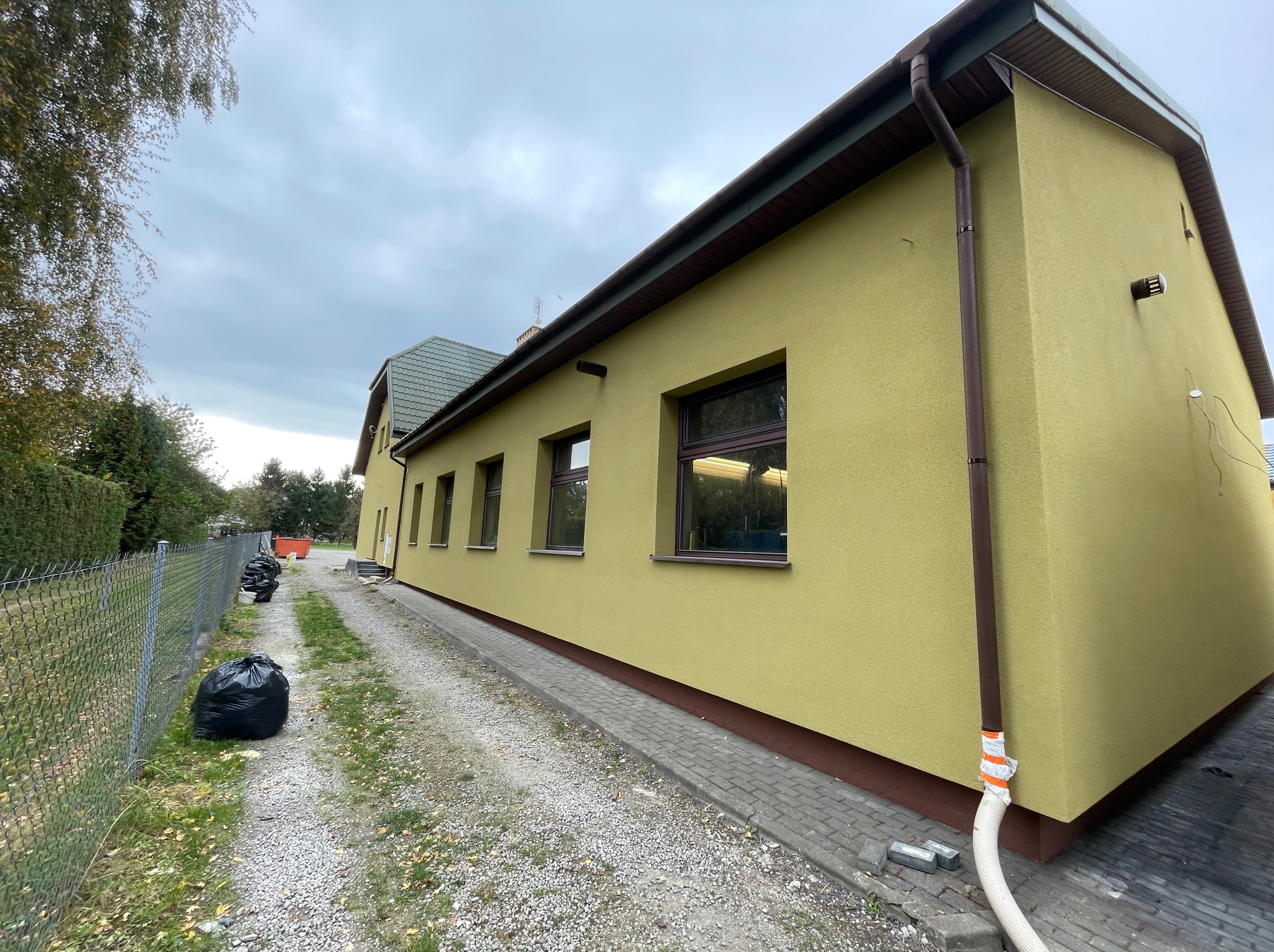 Żółta elewacja budynku z brązowymi oknami i rynnami, widok z boku, z workami na śmieci i wężem na żwirowej ścieżce obok.