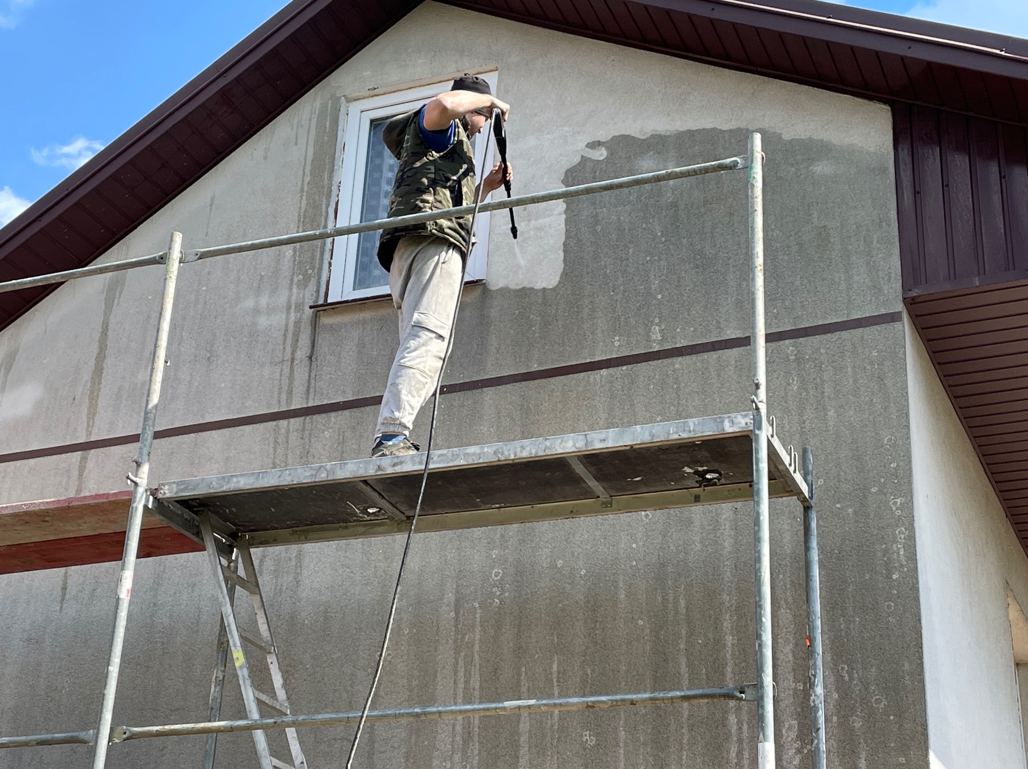 Pracownik na rusztowaniu czyści elewację domu myjką ciśnieniową, widoczny fragment wyczyszczonej ściany, pozostała część jest brudna.