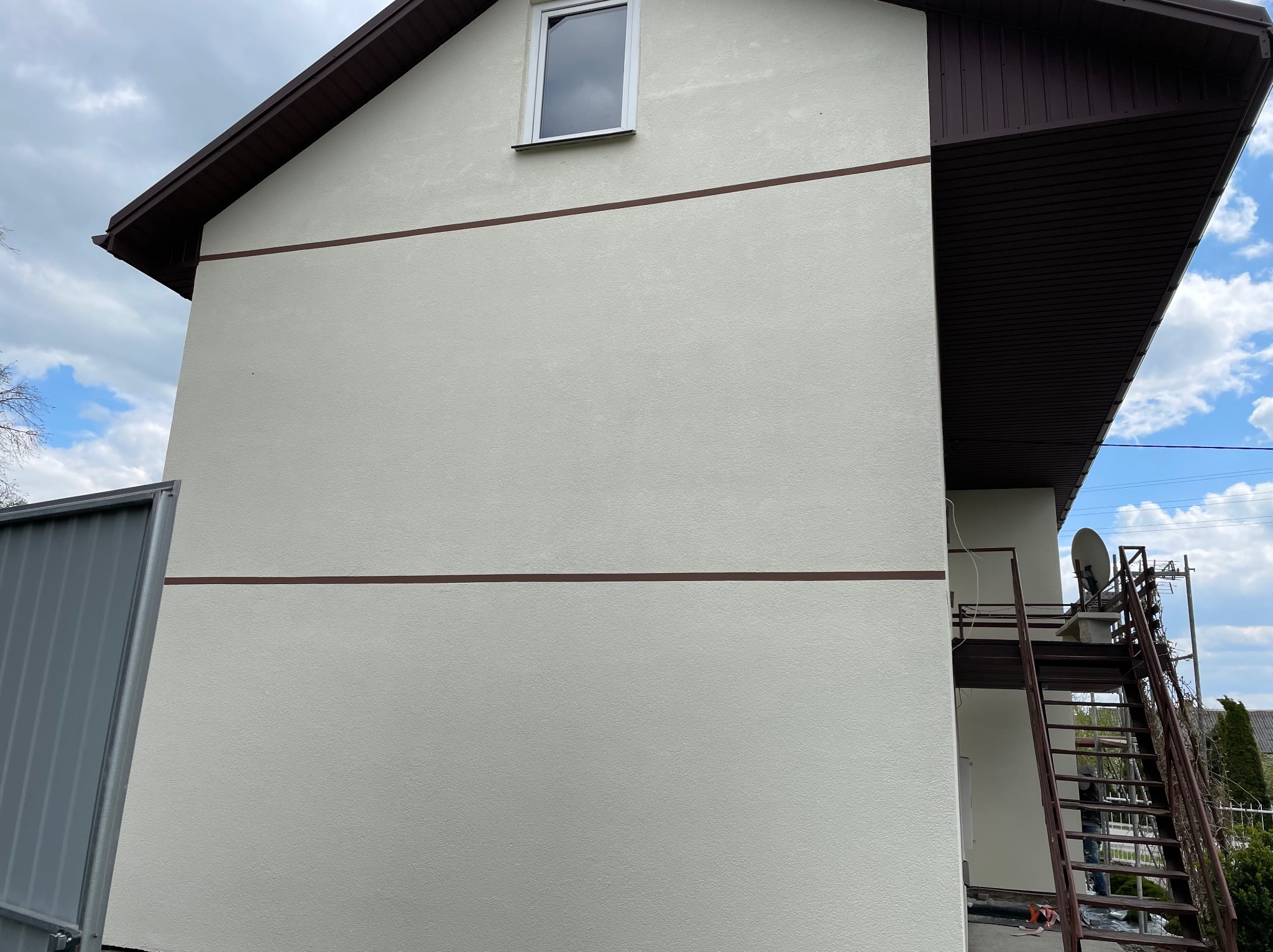 Ściana domu z jasną elewacją i brązowymi akcentami, widoczne schody metalowe prowadzące na dach z anteną satelitarną, pochmurne niebo.