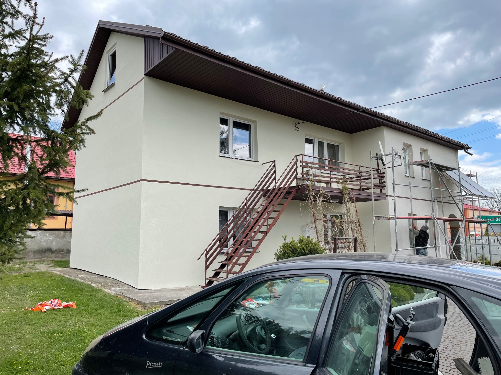 Dwupiętrowy dom po tynkowaniu elewacji, widoczne rusztowania i otwarty samochód z narzędziami przed domem, pochmurne niebo.