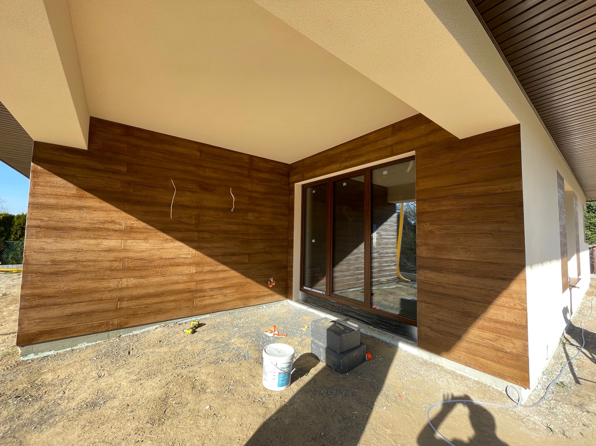 Zadaszony taras z elewacją imitującą drewno, widoczne puszki z farbą i przygotowane podłoże, okno tarasowe z brązową ramą.